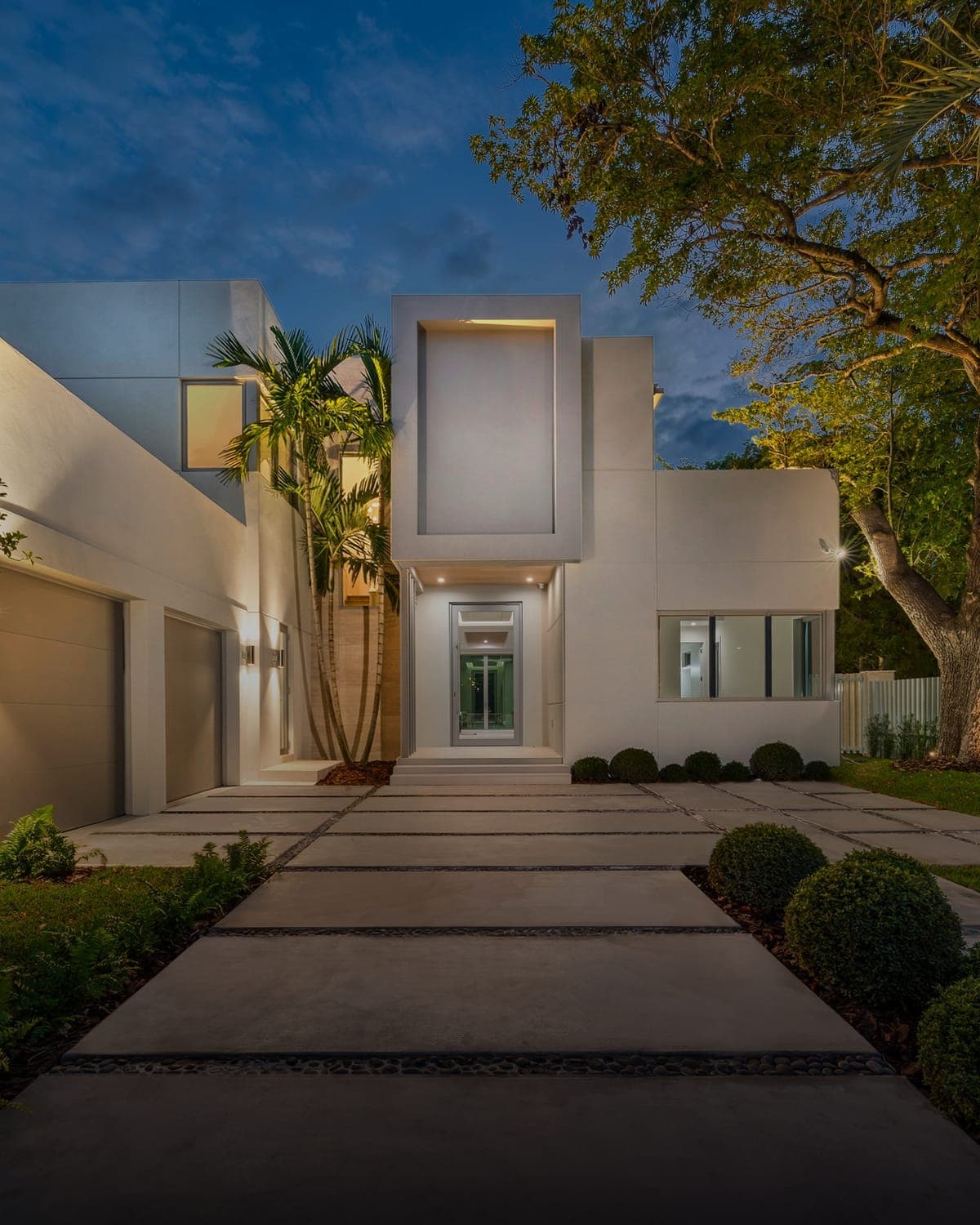 Fachada de residência contemporânea ao entardecer, com iluminação acesa e jardim frontal.